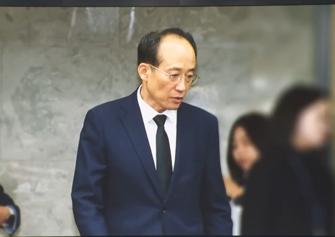 검은 정장 차림의 한 인물이 조용한 표정으로 서 있는 모습